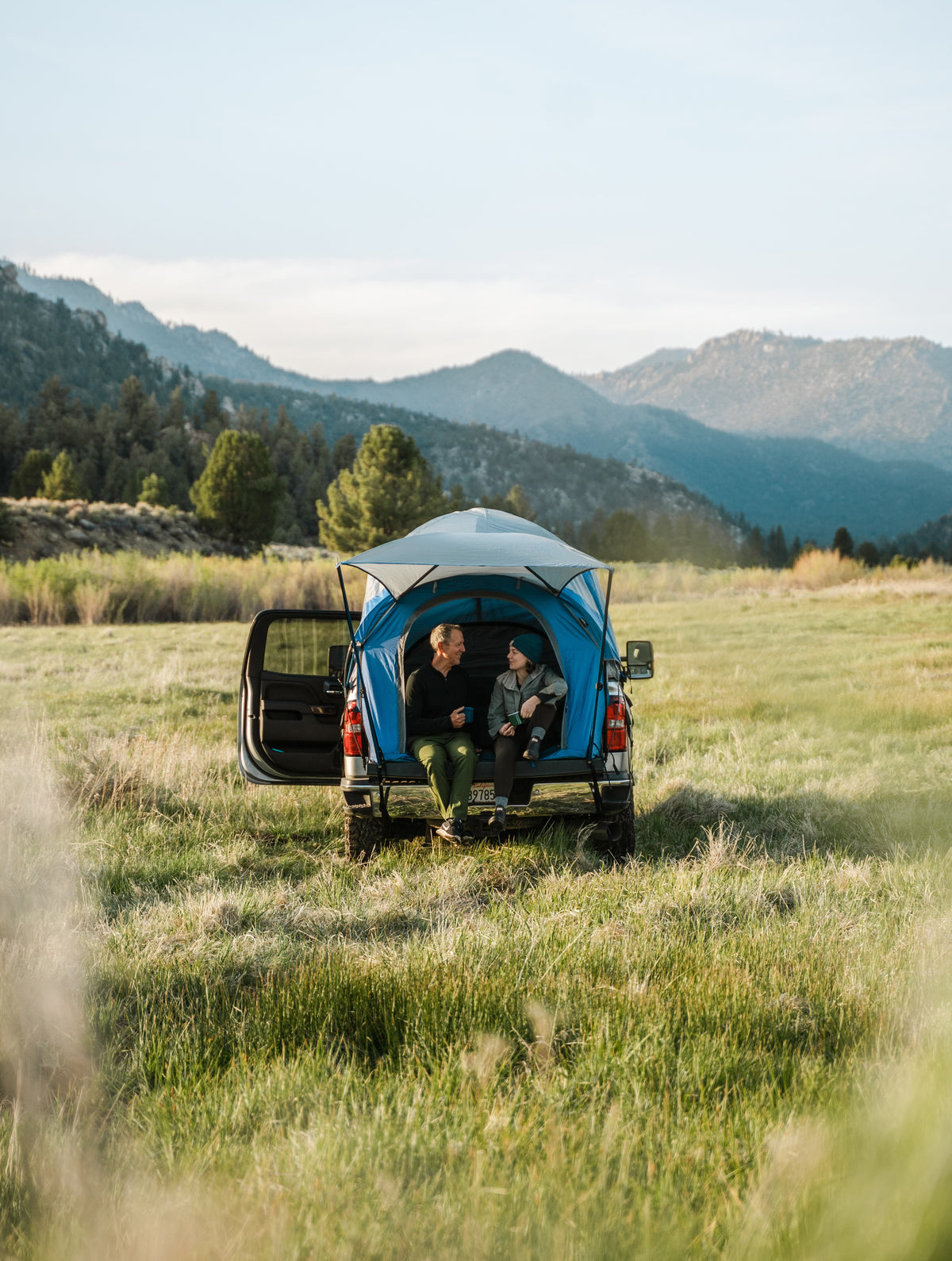 Two people in a truck tent overlanding set up in a field