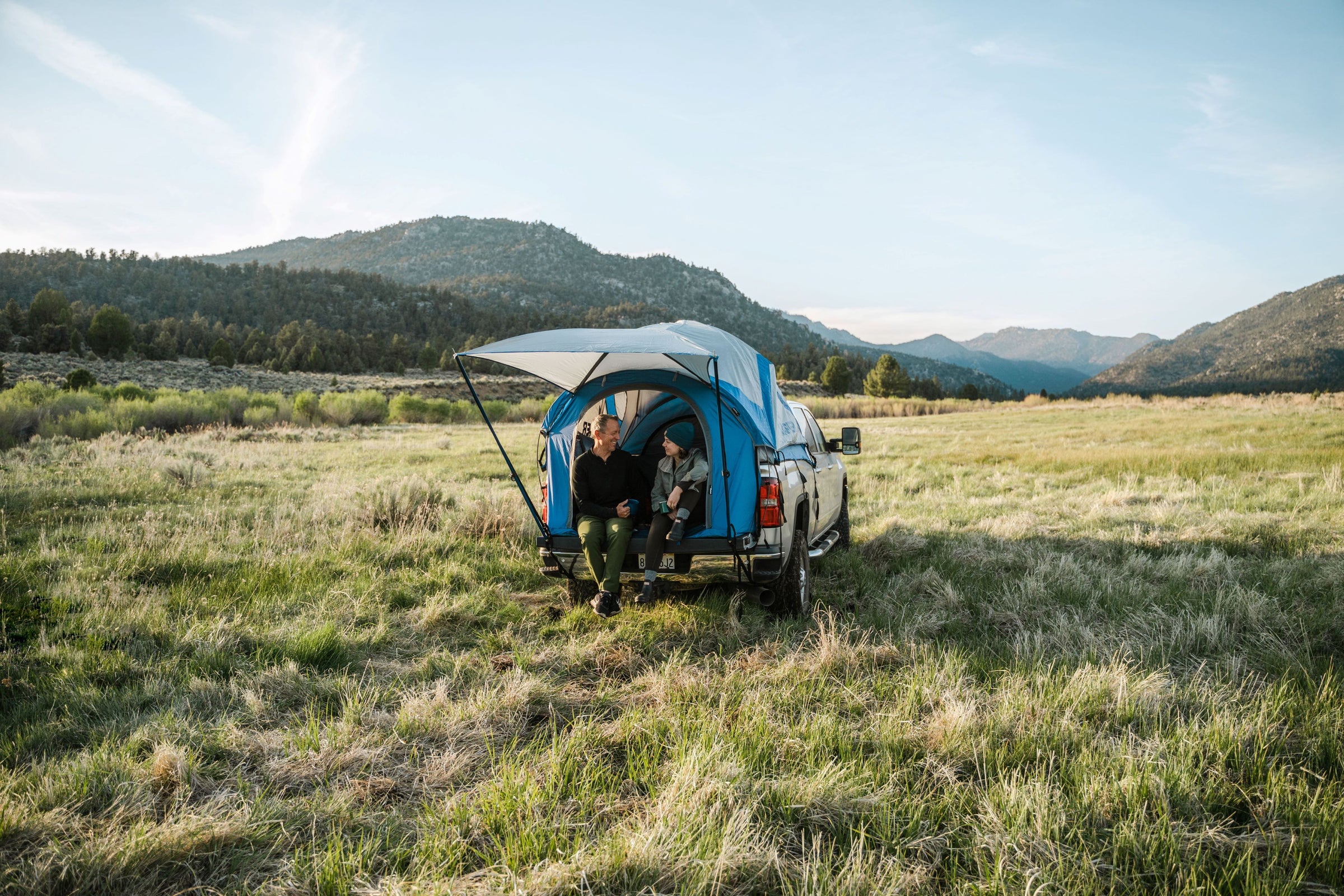 Two people in a truck tent overlanding set up in a field